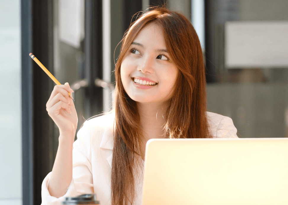 A woman smiling and holding a pencil up in front of a laptop on a desk with a takeaway coffee