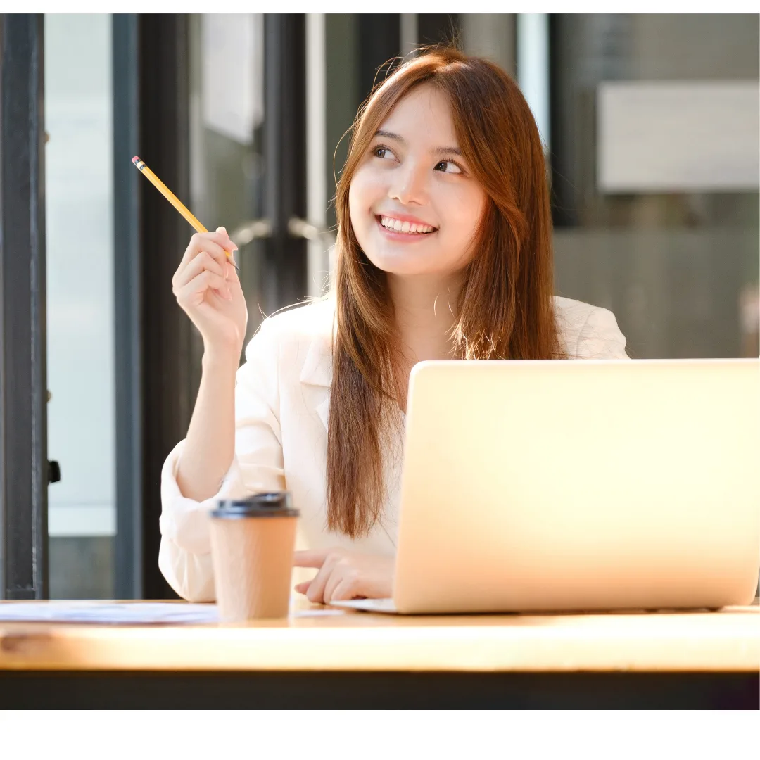 A woman smiling and holding a pencil up in front of a laptop on a desk with a takeaway coffee